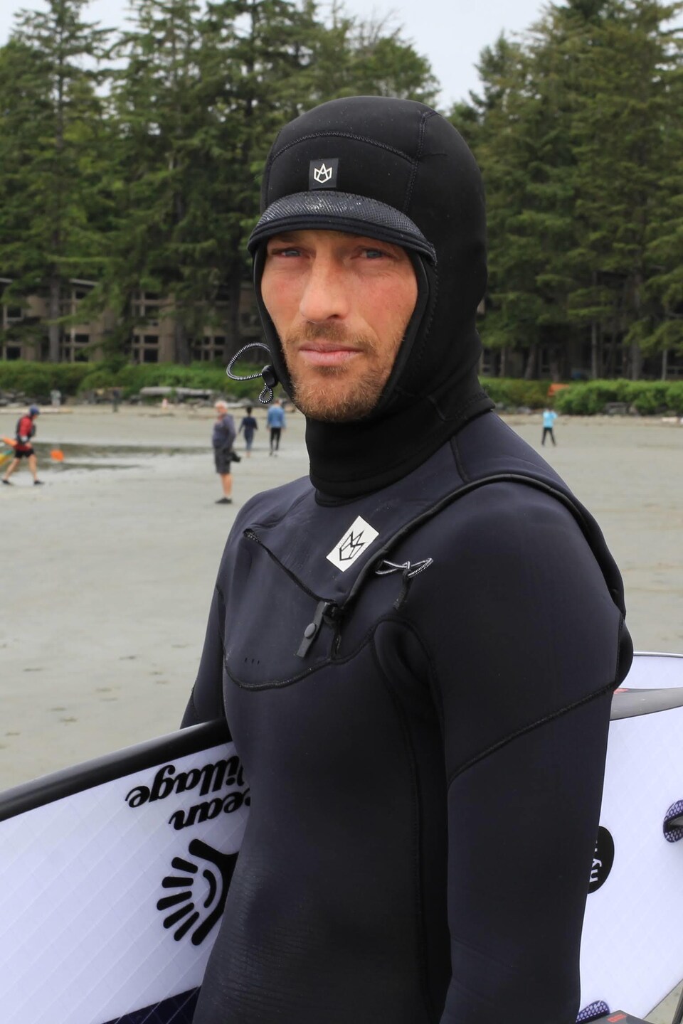 Peter DeVries en combinaison tient sa planche sous le bras, sur une plage.
