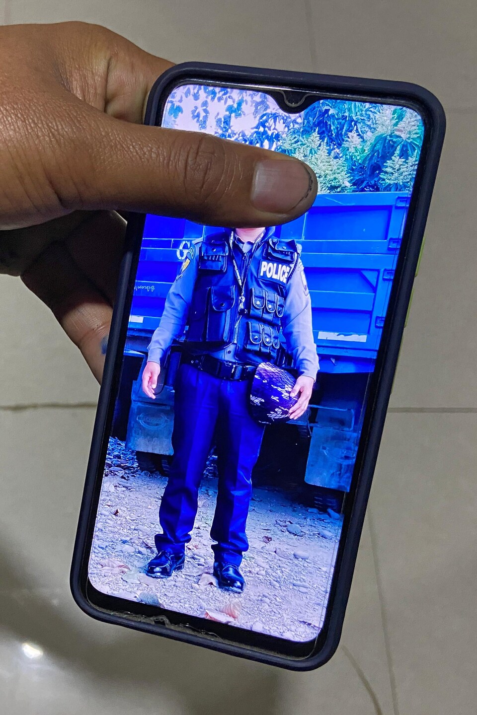 Un homme en uniforme de policier sur une photo montrée sur un téléphone cellulaire. La tête sur la photo est cachée par le pouce de celui qui la montre.