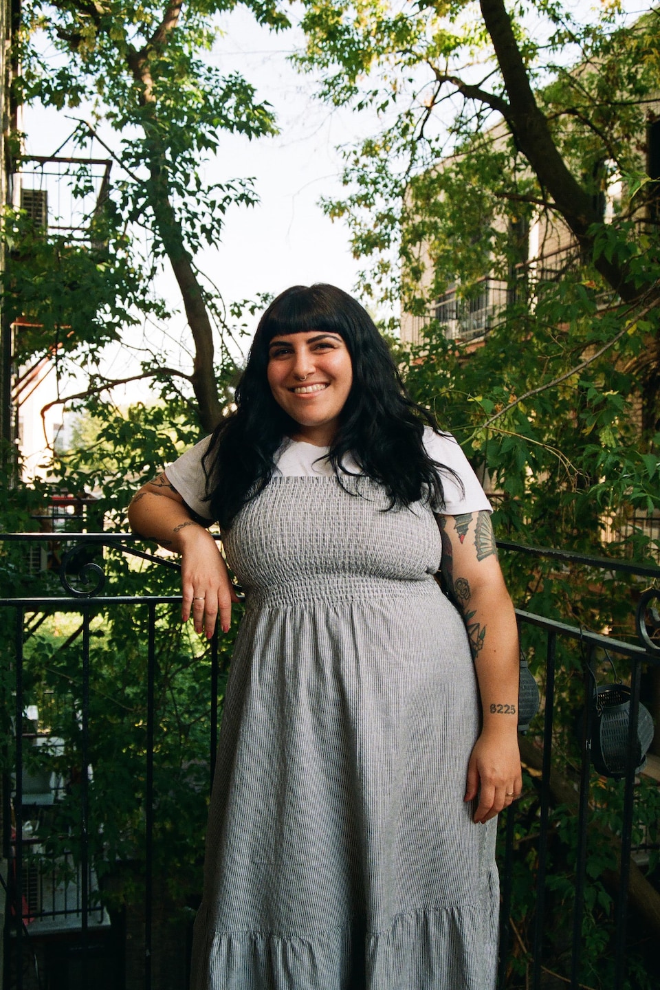 Une femme aux cheveux noirs pose sur un balcon extérieur. Elle est vêtue d'une robe grise et d'un chandail à manches courtes blanc. Elle sourit à pleines dents.