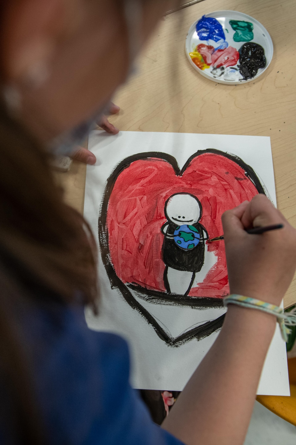 Une enfant peint un grand coeur rouge dans lequel il y a un bonhomme qui tient une planète. 