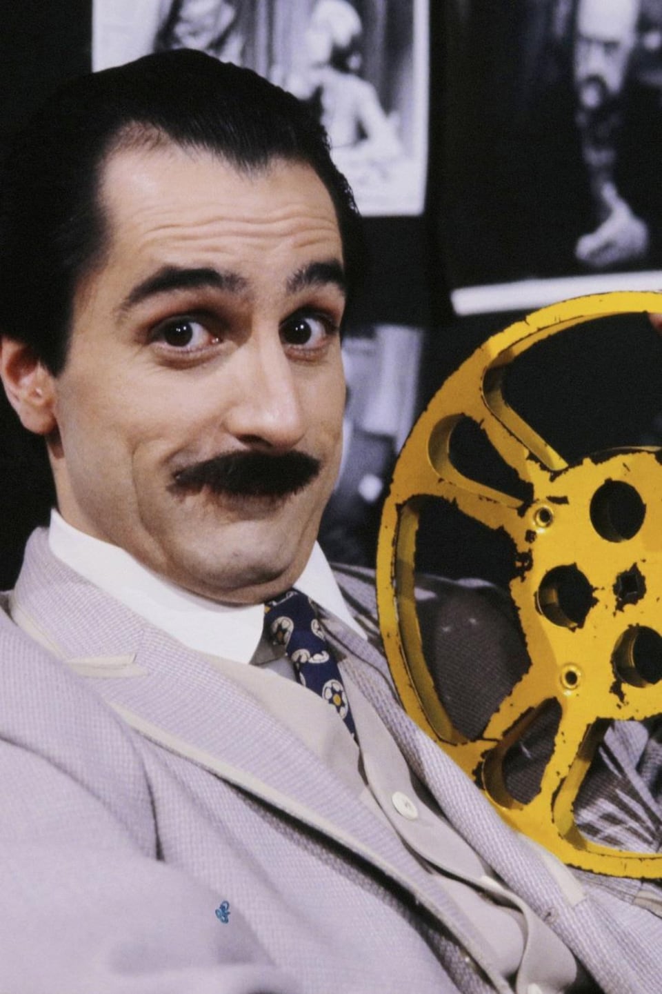 Martin Drainville avec une moustache, tenant dans une main une bobine de film. Derrière lui, des photos de films des années 20.