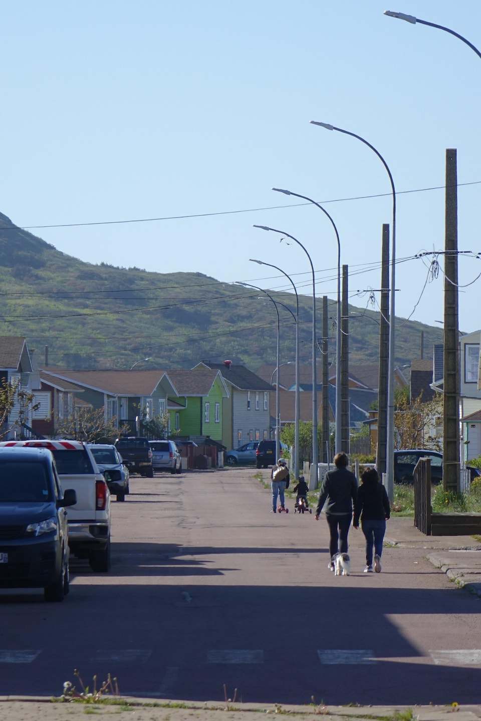 Deux piétons marchent dans la rue d'un village avec un petit mont dans l'arrière-fond. 