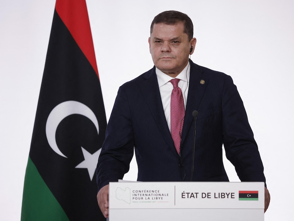 Le premier ministre libyen Abdel Hamid Dbeibah derrière un lutrin et à côté d'un drapeau de la Libye.