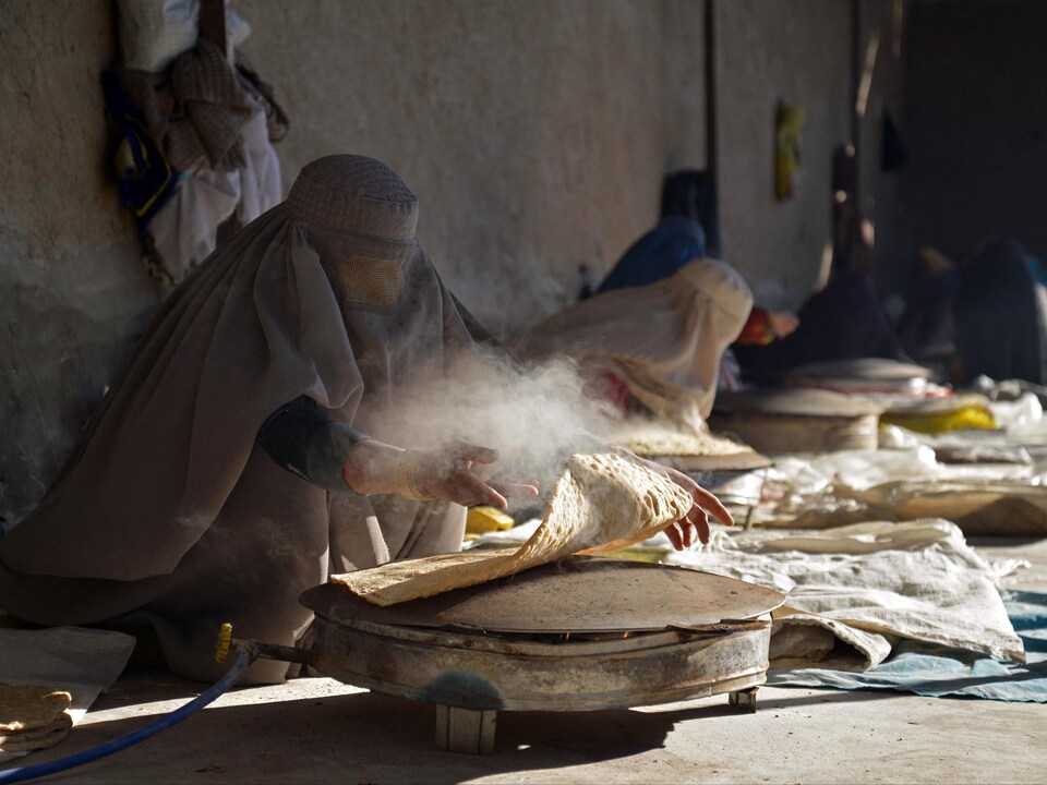 Des femmes en burqa préparent de grands pains plats et ronds sur des fours posés par terre.
