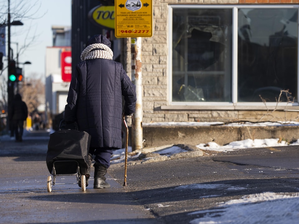 Une personne âgée avec une canne et tirant un chariot marche sur un trottoir à Montréal.
