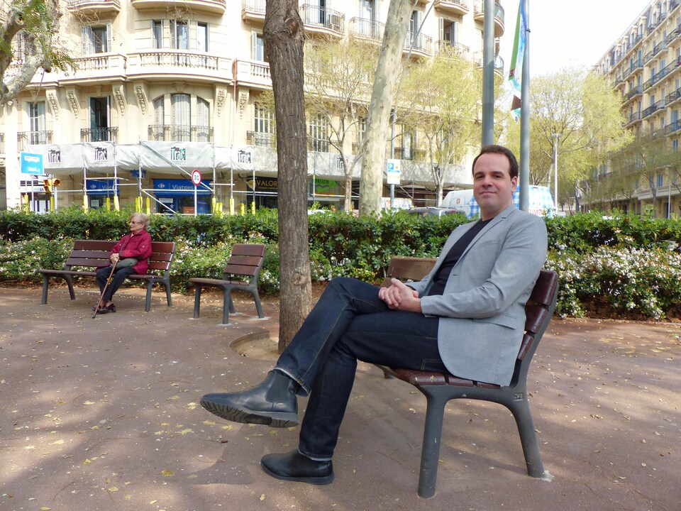L'homme est assis sur un banc dans un parc de Barcelone. Plus loin, une dame seule sur un autre banc.