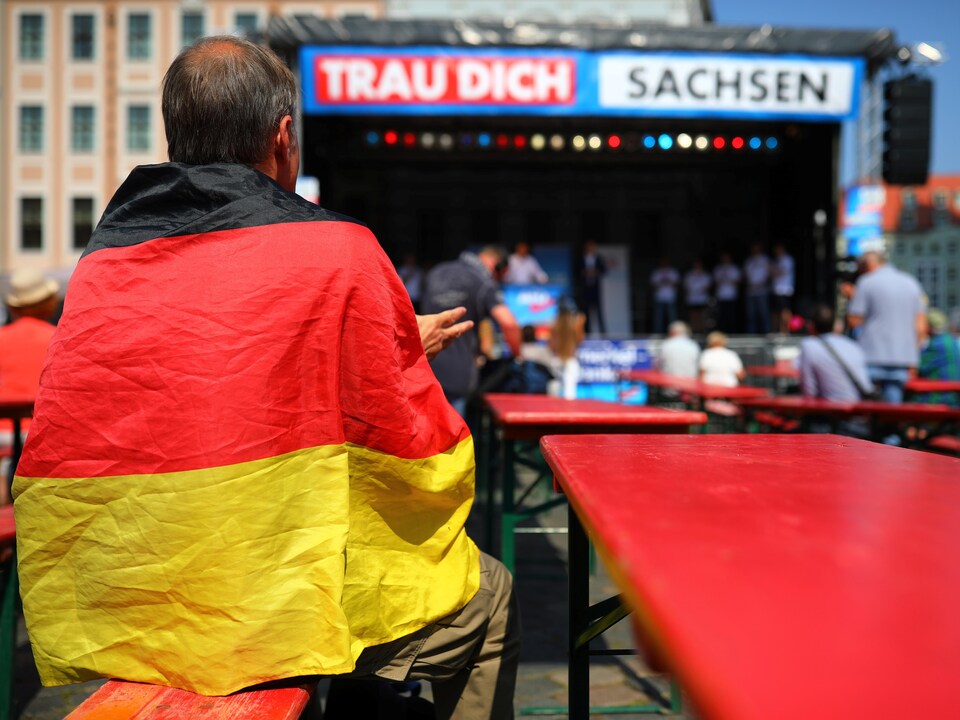 Un homme porte un drapeau allemand sur ses épaules, en regardant des politiciens qui font des discours sur une estrade.  