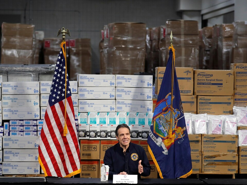 Le gouverneur de l'État de New York parle, assis devant des centaines de boîtes de matériel médical.