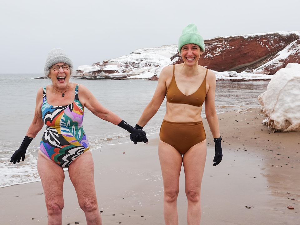 Nicole Grégoire et sa fille Pauline-Gervaise Grégoire se tiennent par la main en souriant, en maillot de bain, sur une plage enneigée des Îles-de-la-Madeleine.