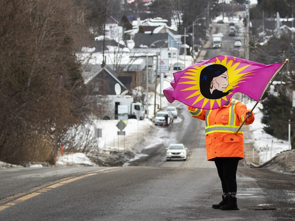 Une femme se tient sur le bord de la route. Elle tient dans sa main gauche un drapeau sur lequel on voit le visage d'une femme autochtone de profil. 