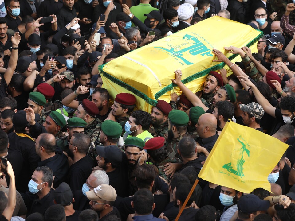 A crowd, including men in military uniforms, carry a flag-covered coffin.