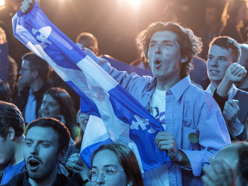 Un jeune homme célèbre parmi la foule. Il tient un drapeau québécois.