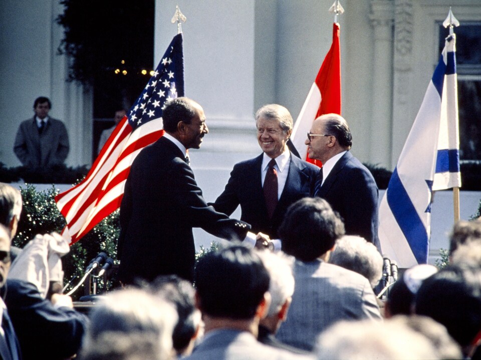 MM. el-Sadate et Begin se serrent la main sur fond de drapeaux des trois États.