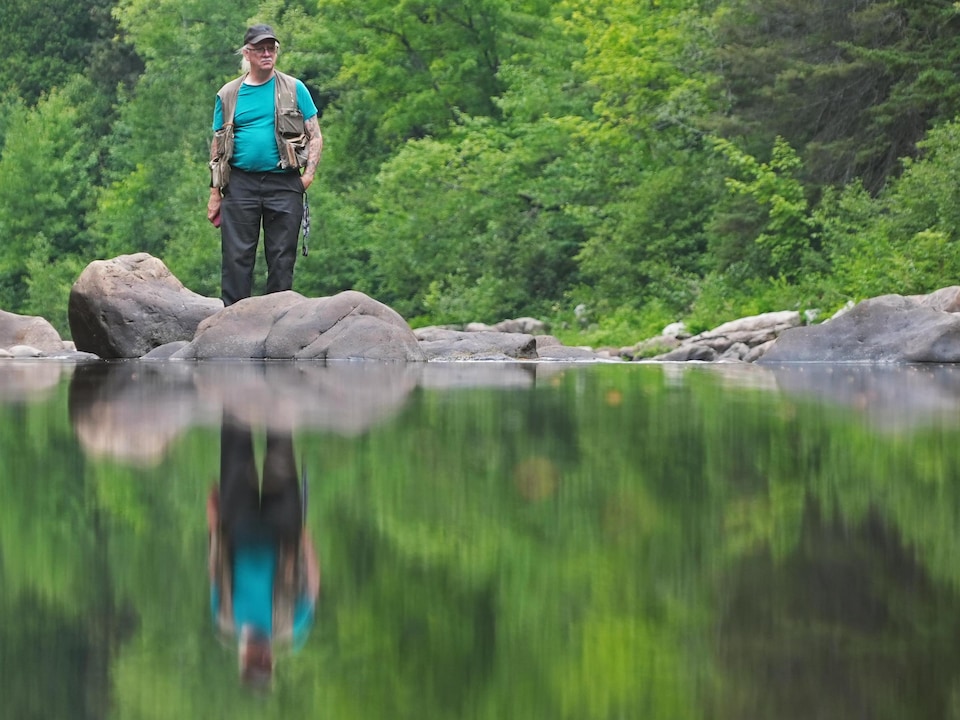Un homme en habit de pêche se tient debout derrière des rochers dans une rivière, calme, entouré de forêt.