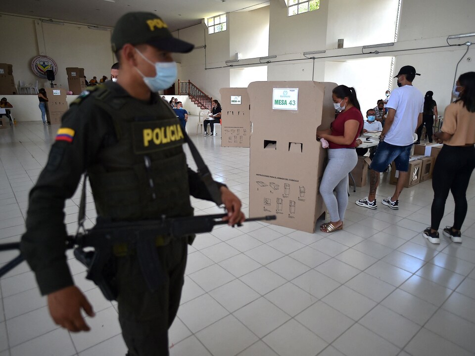 Des électeurs votent tandis qu'un policier armé surveille les environs.