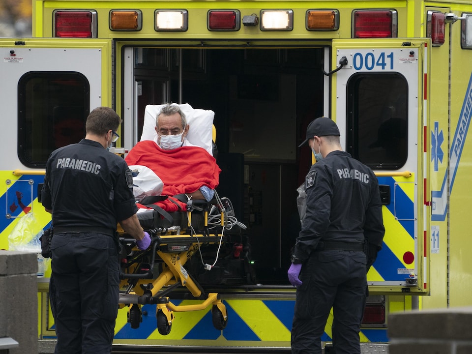 Un homme âgé dans une civière descend d'une ambulance avec l'aide de deux paramédicaux.