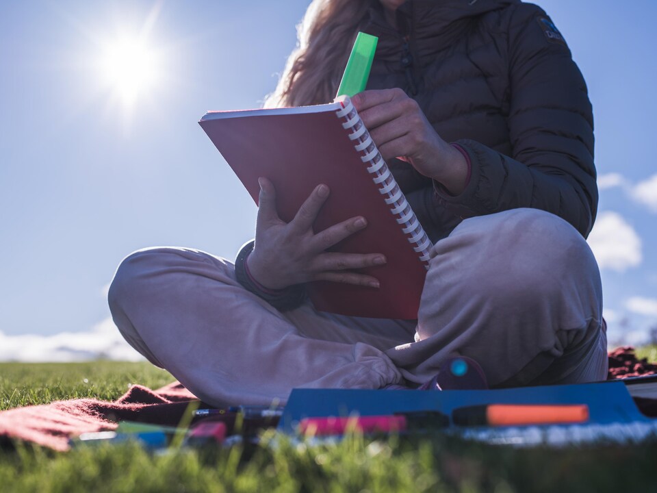 Une élève du secondaire révise ses notes, assise dans l'herbe, sous un soleil éclatant.
