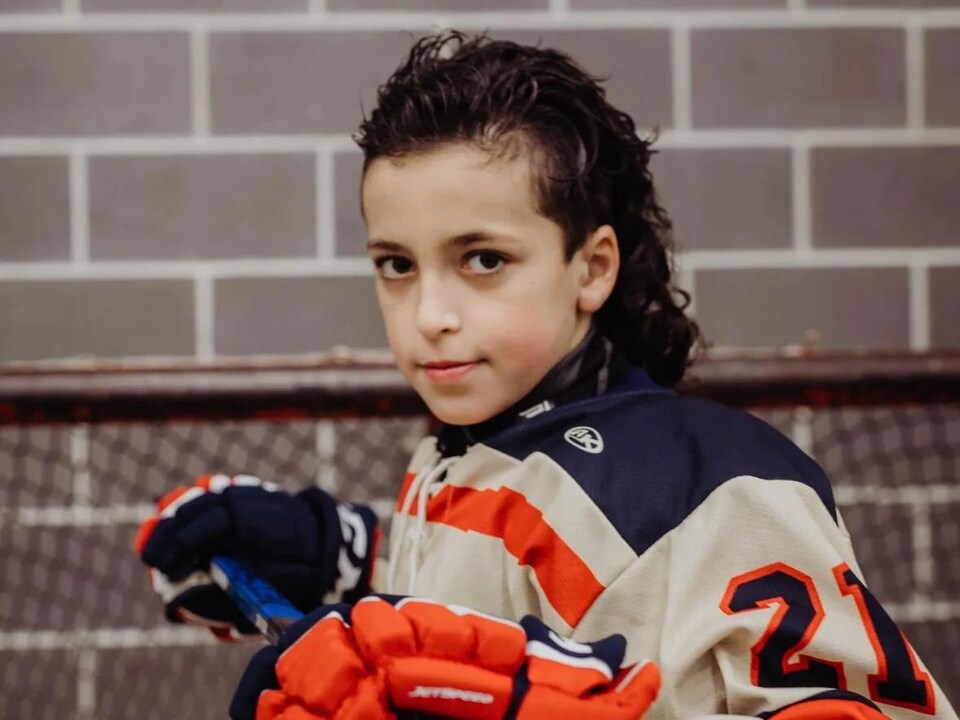 Vêtu de son équipement, un jeune qui arbore une coupe Longueuil pose devant un filet de hockey.