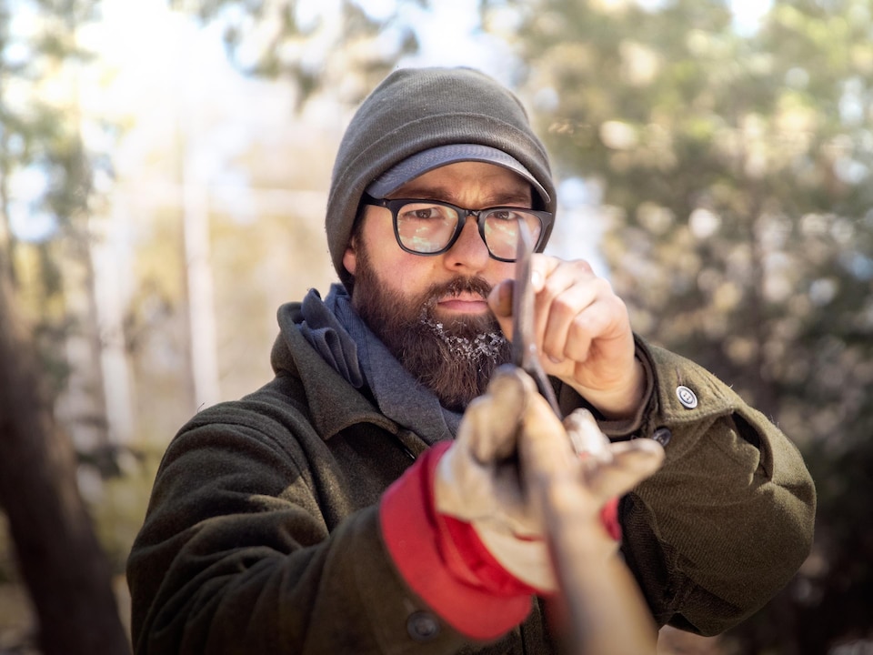 Un homme barbu à lunettes et tuque examine minutieusement la lame d'une hache dans les bois.