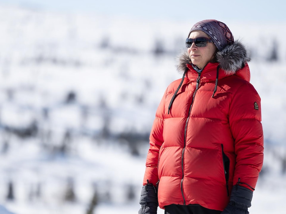 Une femme en manteau d'hiver rouge regarde au loin, devant une montagne enneigée.
