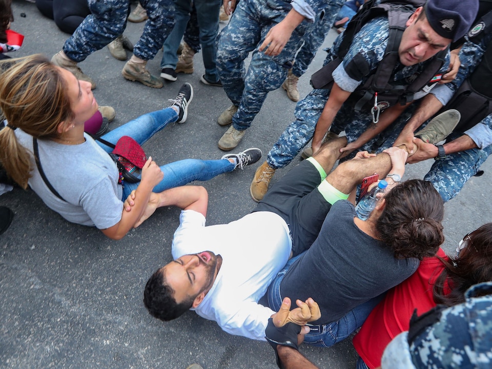 Des personnes portant un uniforme tiennent les jambes d'un manifestant assis sur une route en asphalte. Ce manifestant se tient à d'autres manifestants assis près de lui et une femme retient sa jambe, semblant vouloir empêcher les hommes en uniforme d''emmener le manifestant avec eux.