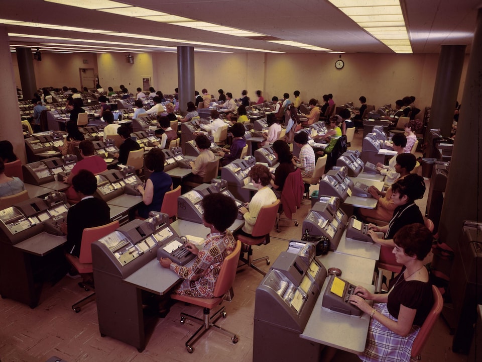 On voit une grande salle où des employées sont assises en rangées en train de taper sur des claviers.