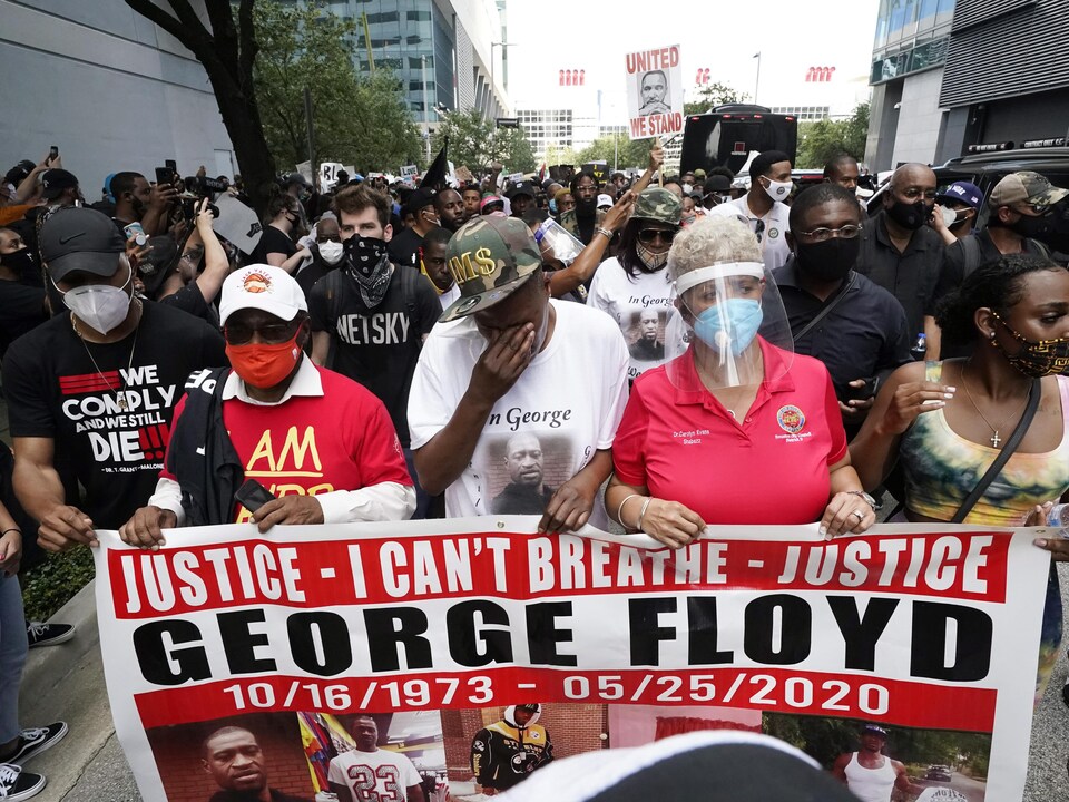 Plusieurs manifestants marchent dans la rue; à l'avant-plan, cinq d'entre eux portent une pancarte réclamant justice pour George Floyd. 