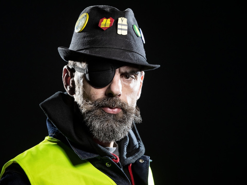 Un homme barbu portant un bandage noir sur son œil droit, un chapeau et un gilet jaune pose pour la caméra.