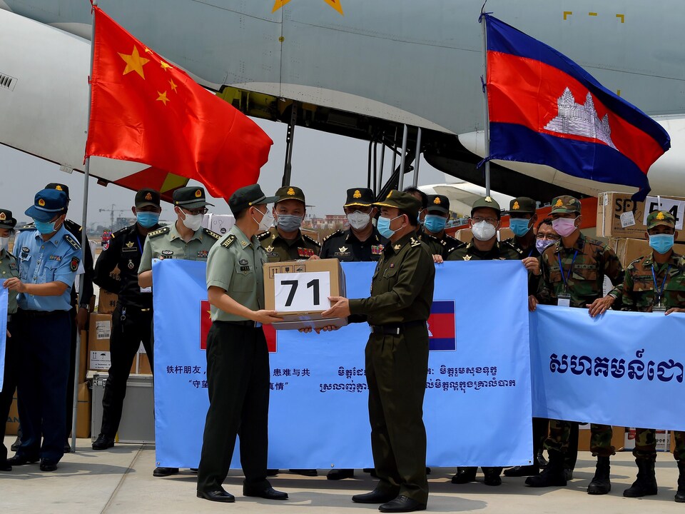 Un homme vêtu d'un uniforme remet une boîte à un autre devant des drapeaux de la Chine et du Cambodge, sur le tarmac de l'aéroport.