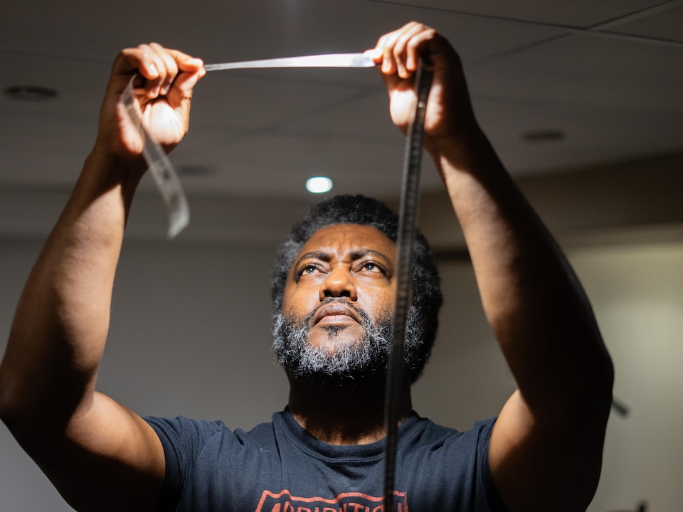 Elyas Tesfaye examine une pellicule tenue entre ses deux mains sous la lumière de son plafonnier, à Winnipeg, en juillet 2026.