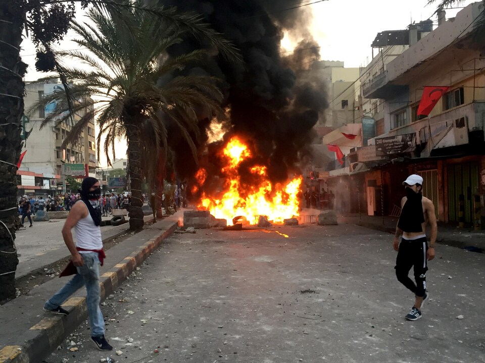 Des manifestants masqués marchent près de pneus en feu dans une rue presque déserte.