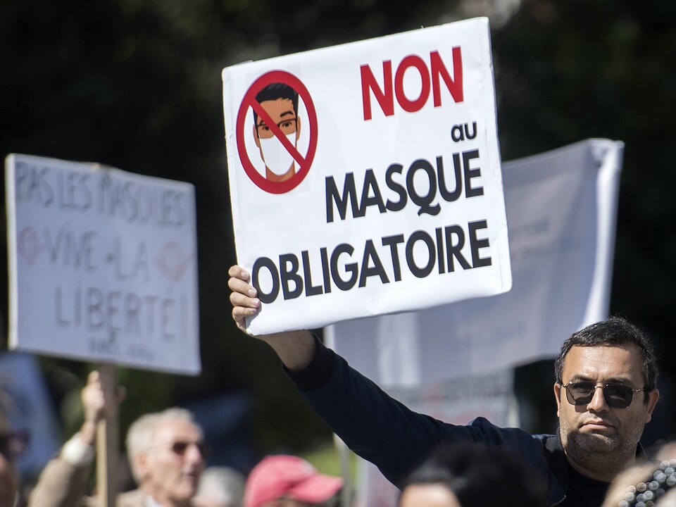 Une foule de personnes, sans masque, manifeste. Un homme tient une pancarte où l'on peut lire : Non au masque obligatoire.