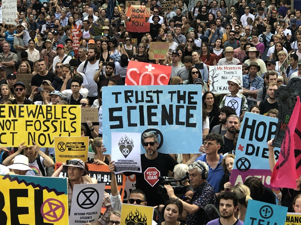Des centaines de gens marchent dans la rue avec des pancartes, dont une sur laquelle il est écrit : Ayez confiance en la science.