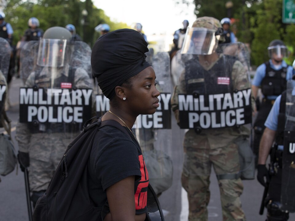 Une jeune femme noire de profile marche devant une ligne de policiers militaires en tenue antiémeute.