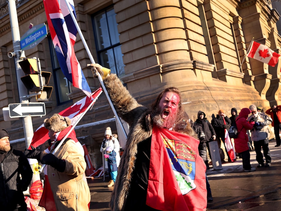 Un homme barbu et maquillé brandit différents drapeaux en criant.