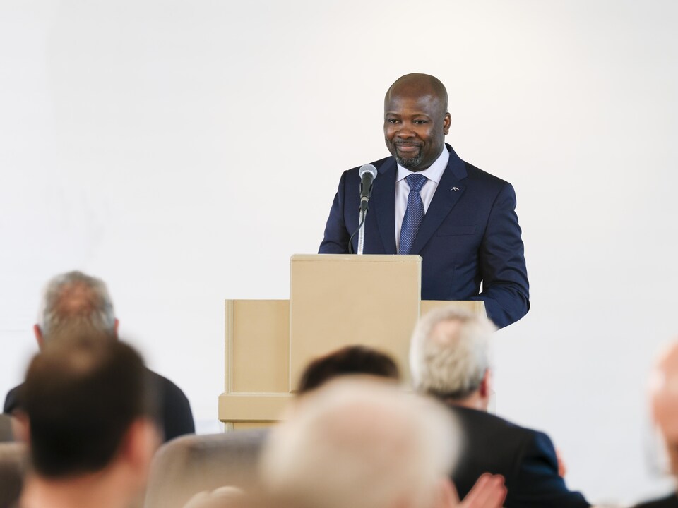 Mapi Mobwano est debout derrière un lutrin lors d'une allocution devant public.