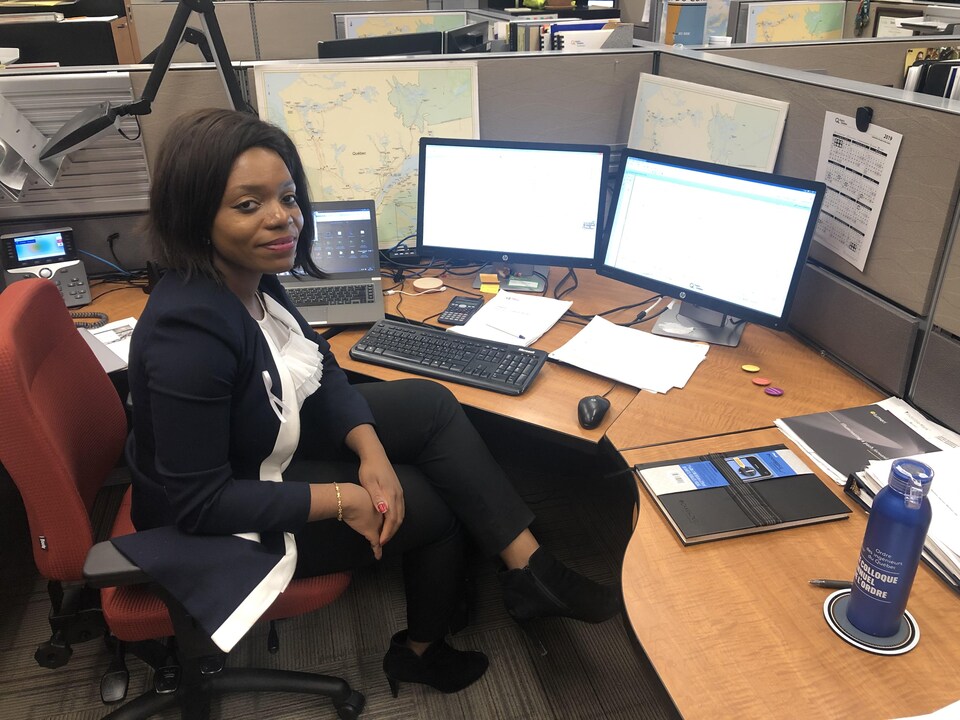Ménélika Békolo est assise à son bureau. Devant elle sont posés des écrans d'ordinateur et un ordinateur portable.