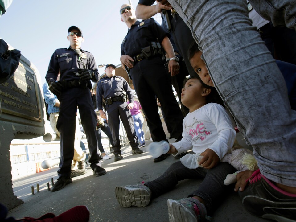 Deux jeunes enfants sont assis au sol, pendant que des gardes-frontières américains sont debout parmi un groupe de migrants. 