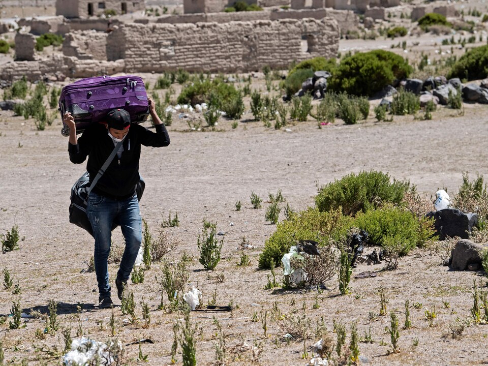 Un homme marche dans une zone désertique avec une valise sur les épaules.
