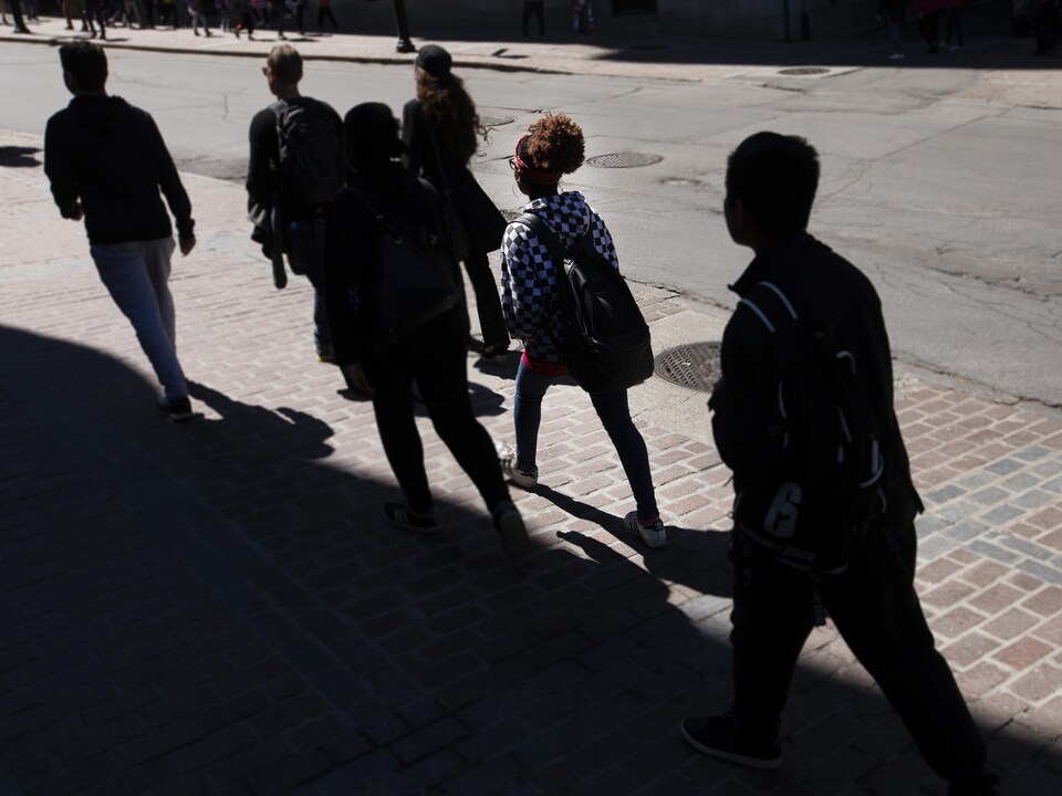 On voit des jeunes, de dos, qui marchent dans une rue du Vieux-Montréal. Ils sont à l'ombre des immeubles.