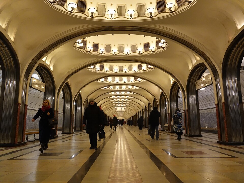 Vue d'ensemble de la station Maïakovskaya, avec sa grande allée centrale, ses coupoles éclairées au plafond et son plancher carrelé de marbre, ainsi que les quais de part et d'autre.