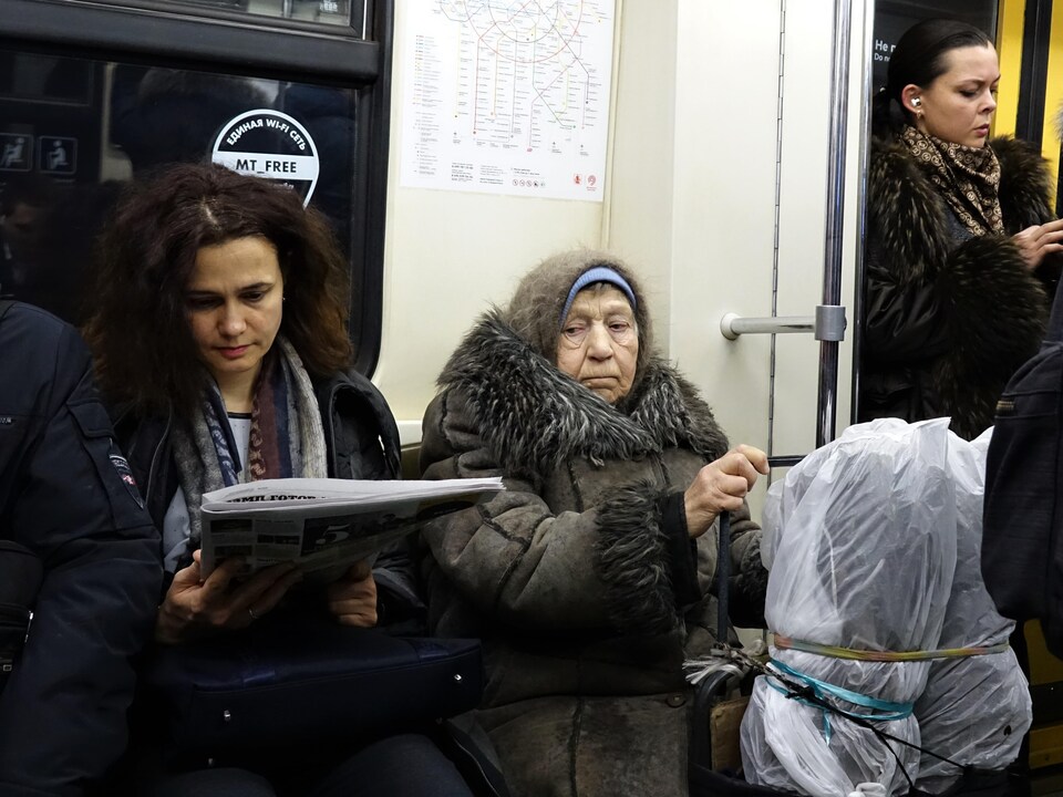 À l'heure de pointe dans le métro de Moscou, on voit une dame âgée et ses bagages enveloppés de plastique à côté d'une jeune femme en train de lire le journal.