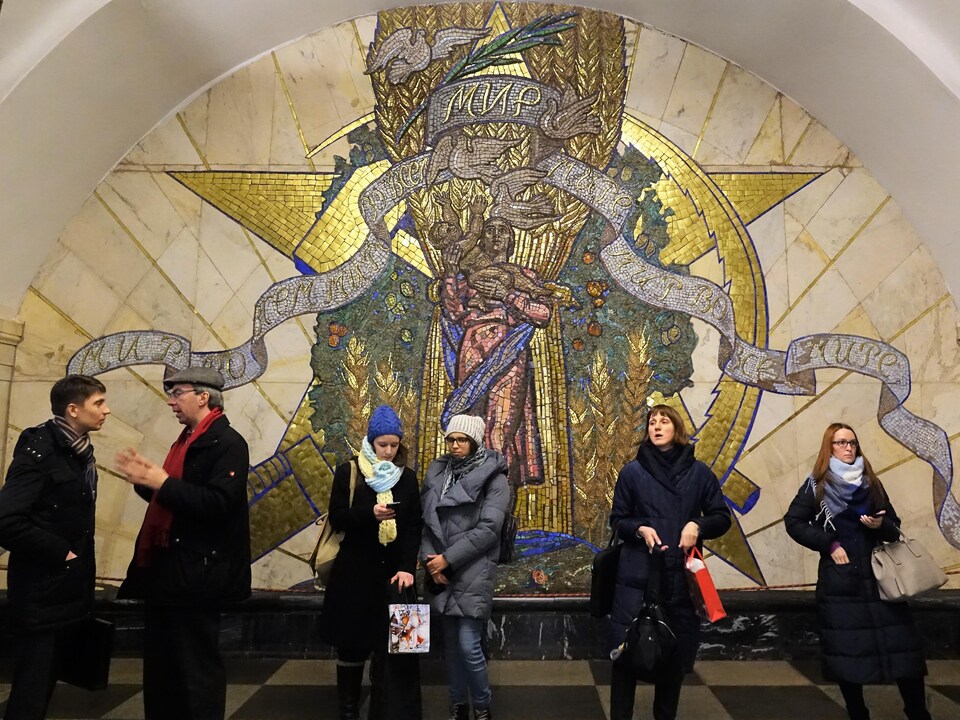 Des gens attendent, discutent et consultent leur téléphone, devant une fresque représentant une femme et un enfant qui tend les bras vers des colombes, à la station Novoslobodskaya.