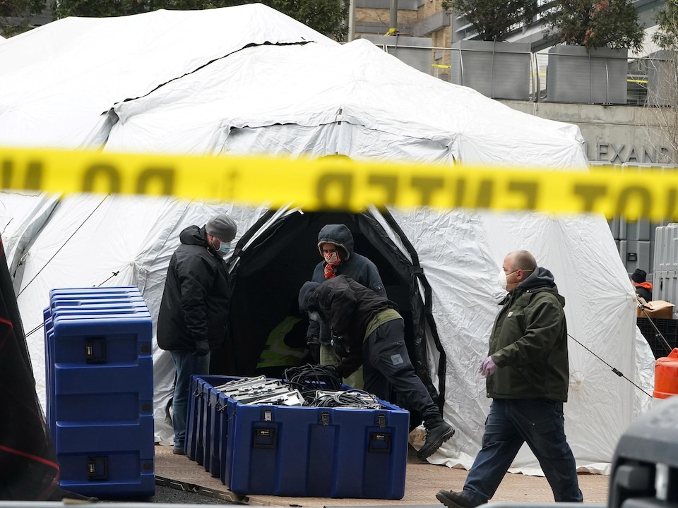 Men wearing masks carry equipment in a white tent.  A ribbon limits access to the site.