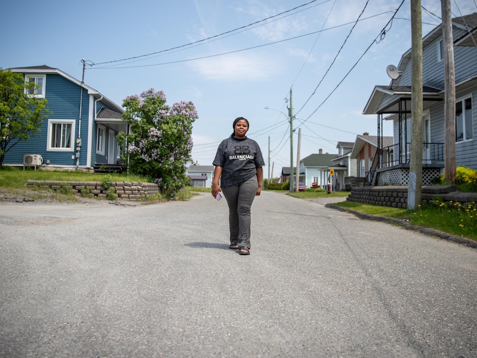 Une jeune femme vêtue d'un t-shirt gris avance au milieu d'une rue résidentielle bordée de maisons.