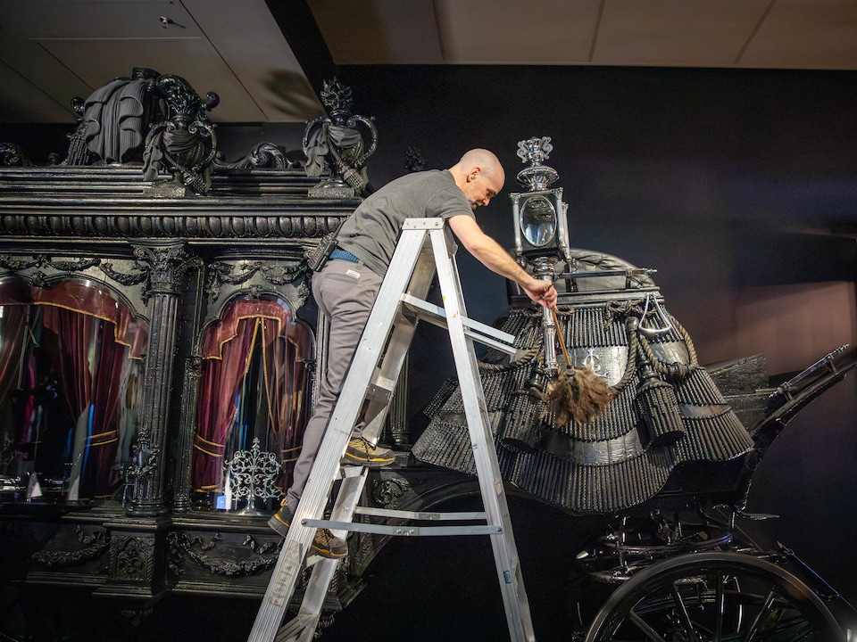 Un employé du Musée de la civilisation, debout sur un escabeau, utilise un plumeau pour nettoyer les éléments décoratifs à l'arrière d'un imposant corbillard hippomobile noir.