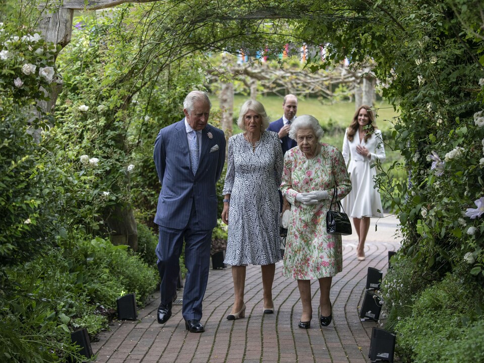 Le prince Charles, Camilla et la reine Élisabeth II passent sous une arche végétale dans un parc.