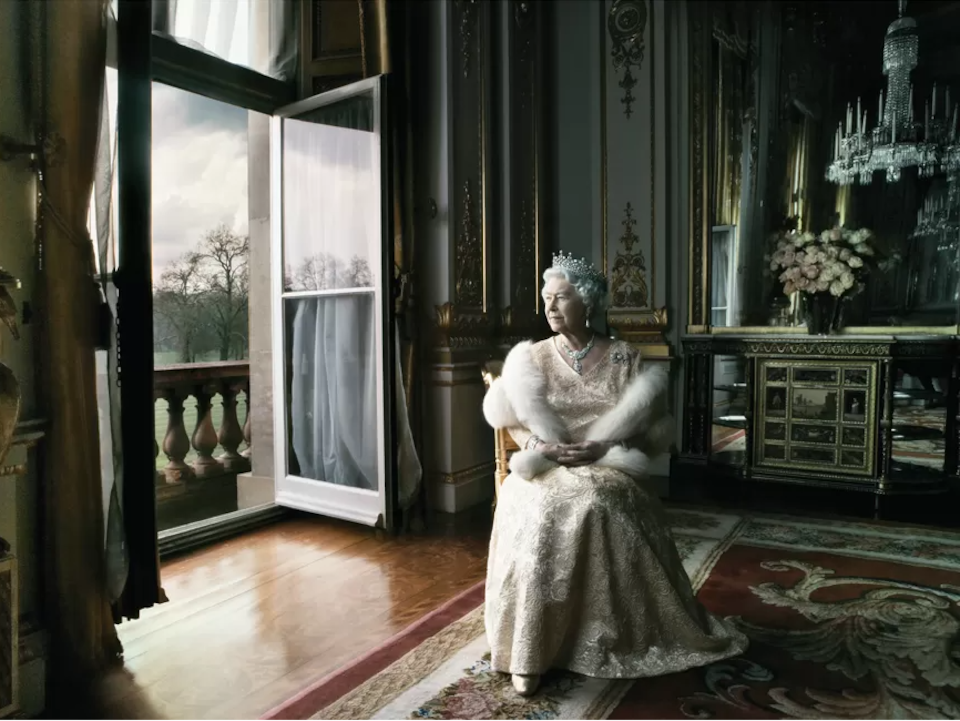 La reine drapée de vêtements royaux regarde vers sa droite, assise dans une pièce dont la fenêtre ouvre sur un balcon. 