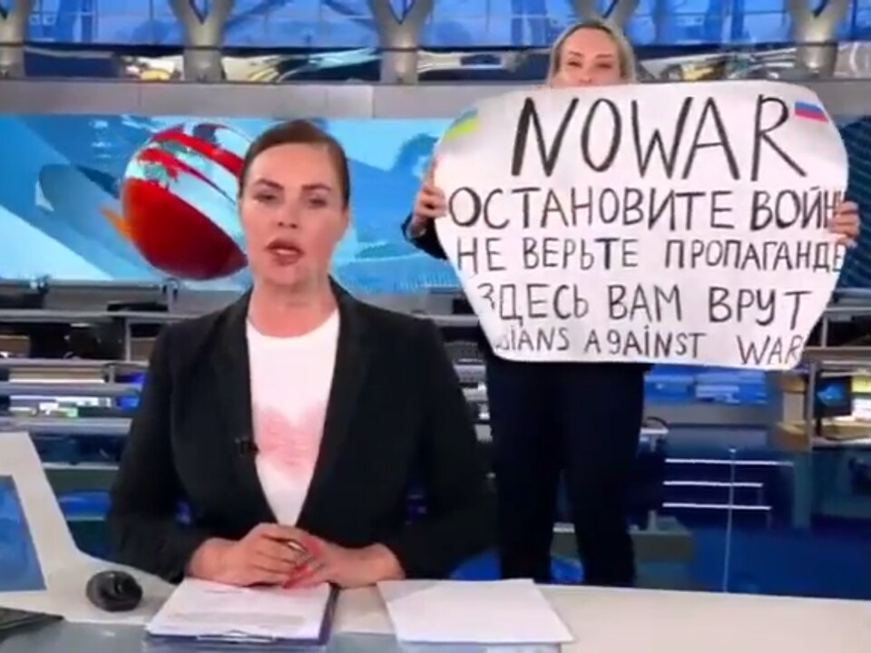 Une femme, qui a surgi sur un plateau de télévision, tient une pancarte disant « Non à la guerre. Ne croyez pas la propagande. On vous ment, ici », derrière une lectrice de nouvelles. 