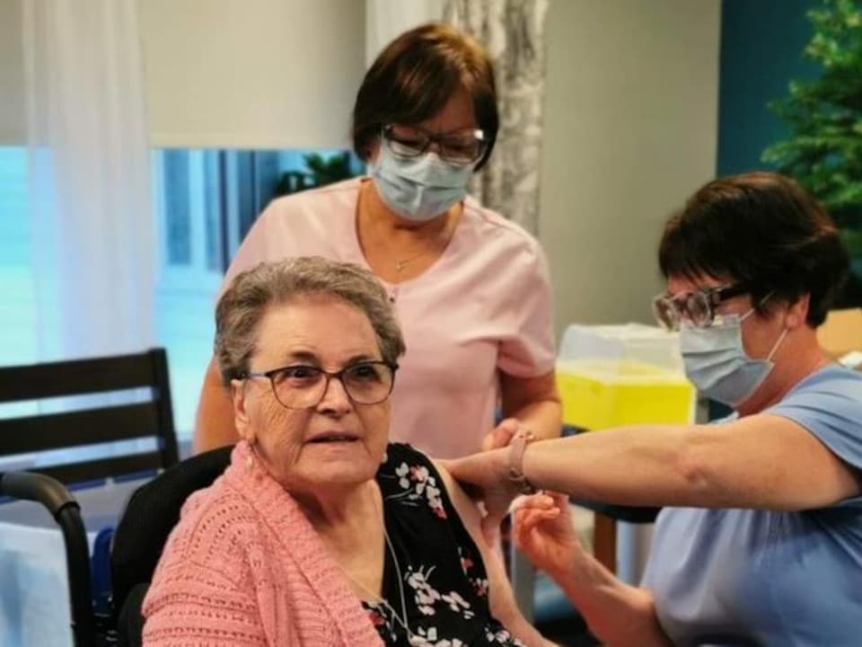 Une dame de 84 ans reçoit le vaccin entourée de deux infirmières.
